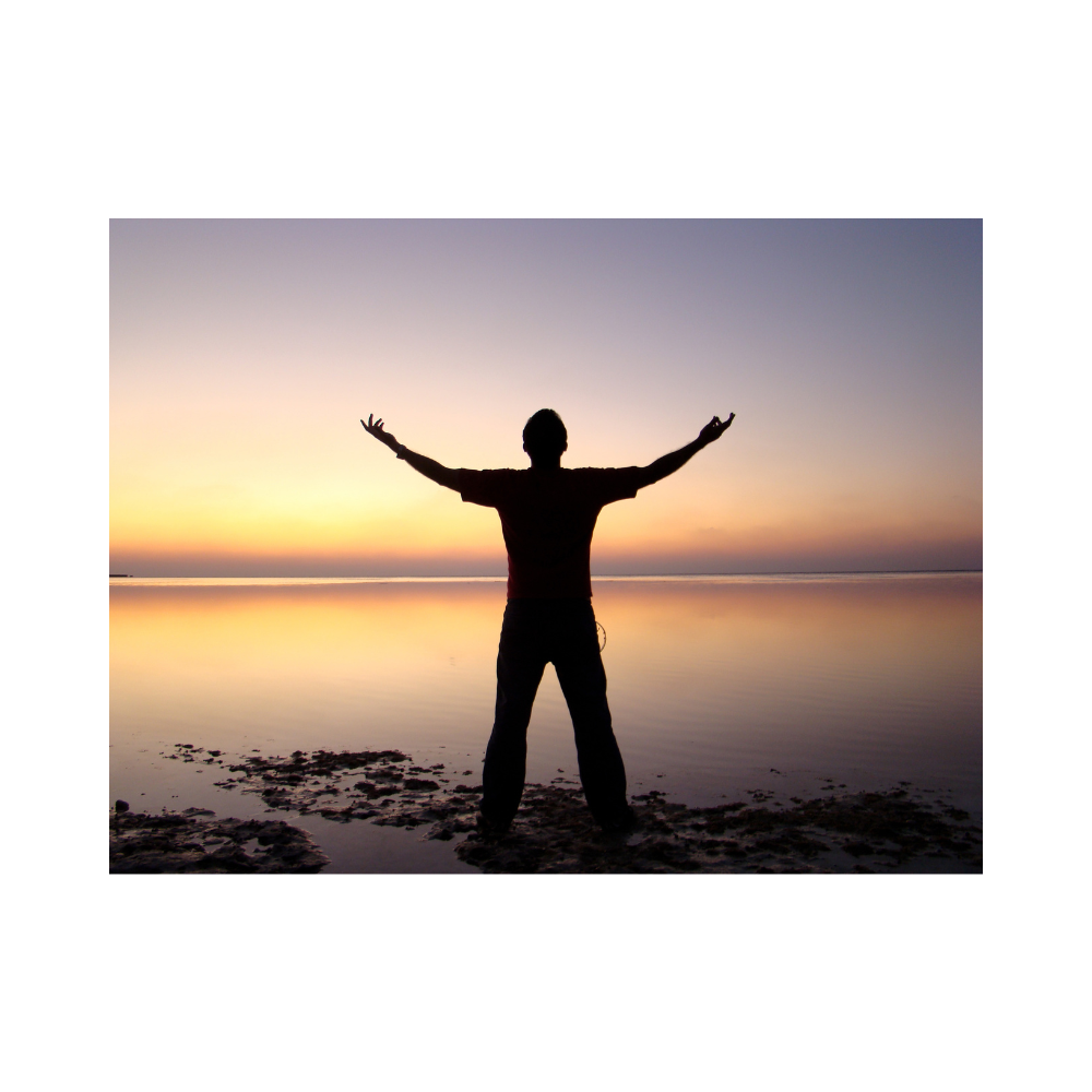 Man on a beach facing the water at sunset in a pose of surrender - arms open wide, face looking up to the sky